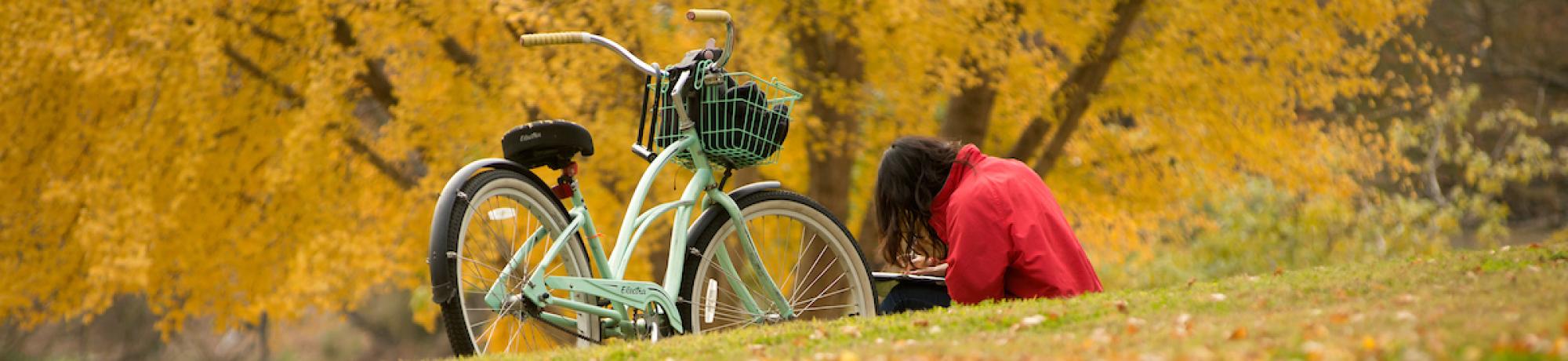 Student reading in leaf-covered grass, next to bike.